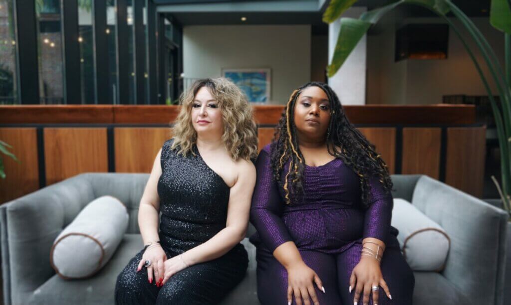 Two women sit on a modern-style gray velvet sofa in a lounge. Windows illuminates them from the left. There is green foliage on either side of the frame. On the left, Lubana has light skin and medium blonde shoulder-length wavy hair, wearing a sparkly black dress that is sleeveless and off the shoulder on the left side. On the right, Andrea has medium-dark skin and long black hair in twists with gold highlights, wearing a sparkly royal purple jumpsuit. Both women are slightly smirking at the camera and have a calm demeanor with their hands resting on their legs.