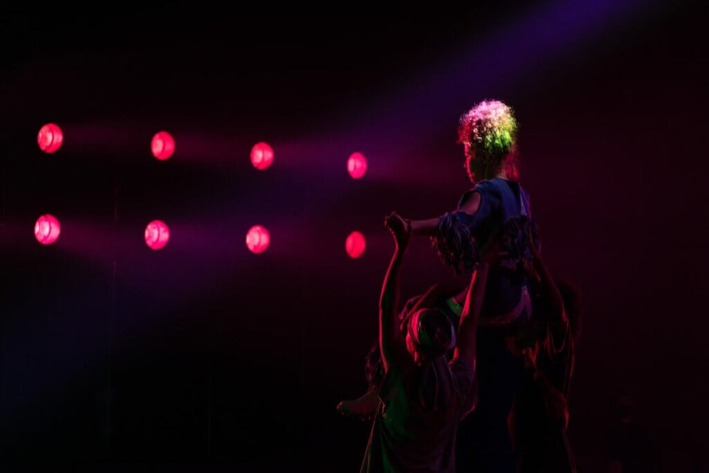 A light-skinned curly-haired haired femme in denim fringe is hoisted up onto the shoulders of another Black femme by two other Black femme performers -- one in a du-rag, one with her hair out in a soft afro. A row of pink lights shine on their faces, and they are backlit in an otherwise dark scene.	