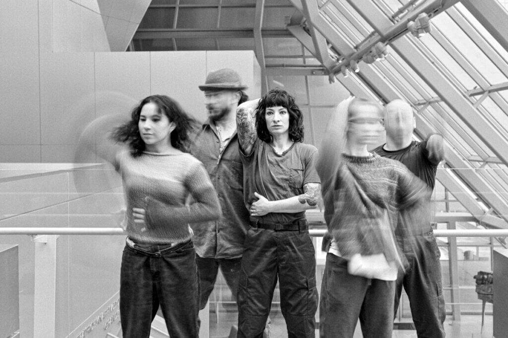 A black-and-white photo of five individuals in an indoor space with large windows. The person in the center, a woman with tattoos and short dark hair, stands still with one arm raised, looking directly at the camera. The other four individuals, two on each side, are in motion, creating a blurred effect. One person wears a hat, another has long hair, and the others appear to be moving their arms or bodies rapidly. The overall composition captures a dynamic and artistic moment with a mix of stillness and motion.