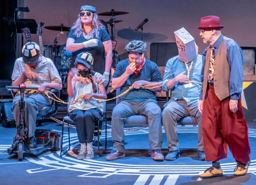 A theatrical performance featuring six actors on stage. Five of the actors are seated in a row, each with unique props and costumes. One actor wears a paper bag labeled "CENNET" over their head, another is in a mobility scooter, and another has a black plastic bag on their head. They are all holding a thick yellow rope. A standing actor, wearing a red hat, blue shirt, patterned tie, and red trousers, appears to be leading or addressing the group. A female flight attendant is standing behind the seated actors wearing sunglasses, gloves, and a cap with patches. The backdrop includes musical instruments, suggesting a lively and dynamic scene.