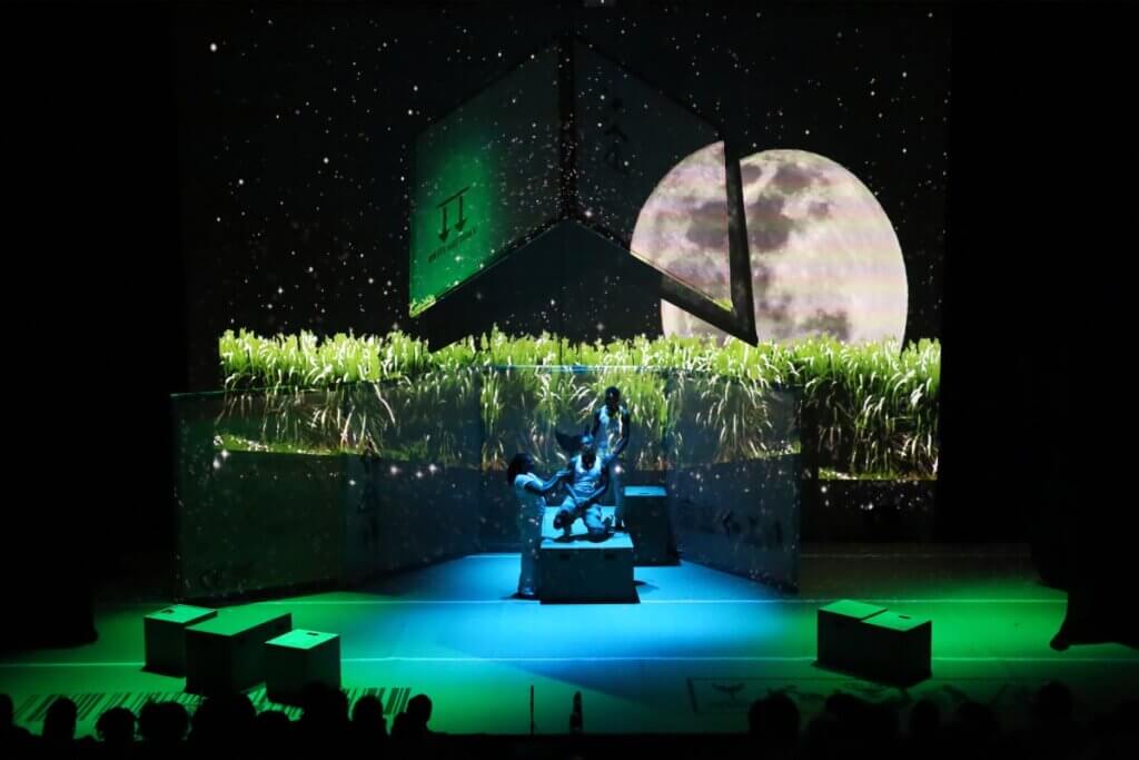Three members of the Yankee Bajan ensemble, Black actors of Barbadian descent, dance beneath a green projected landscape of sugar cane fields in Barbados WI. Three actors, one kneeling on an elevated box, move beneath the large pie-shaped moon, as packing boxes and a cardboard floor are seen downstage of the image. An A-Frame projection screen is flown overhead and reflects the images as well.