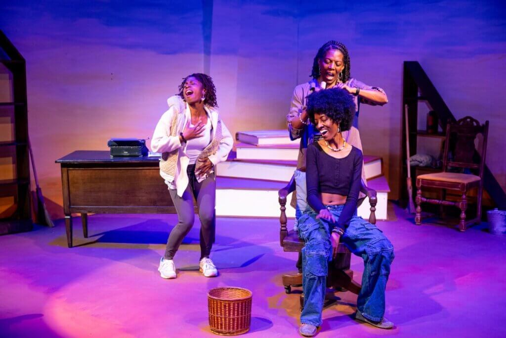 A dark-skinned woman wearing twists is singing and dancing. An older dark-skinned woman with braids sings while parting the hair of another dark-skinned woman who is sitting in a chair and laughing. The stage has a wooden desk with a typewriter on it and bookshelves.