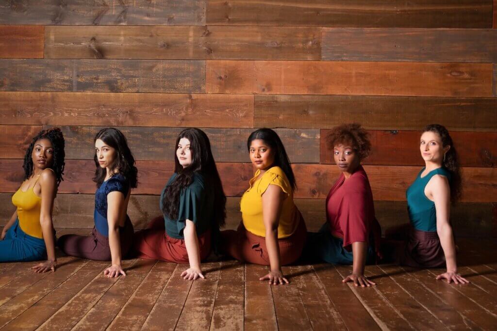 Six women sit in a horizontal line on the floor with their left palms firmly planted on the ground while looking directly at the camera over their left shoulders. The floor and the wall behind them are dark wood panelling. They are dressed in various pants and tops in hues of marigold, burgundy, royal blue, turquoise, and plum.