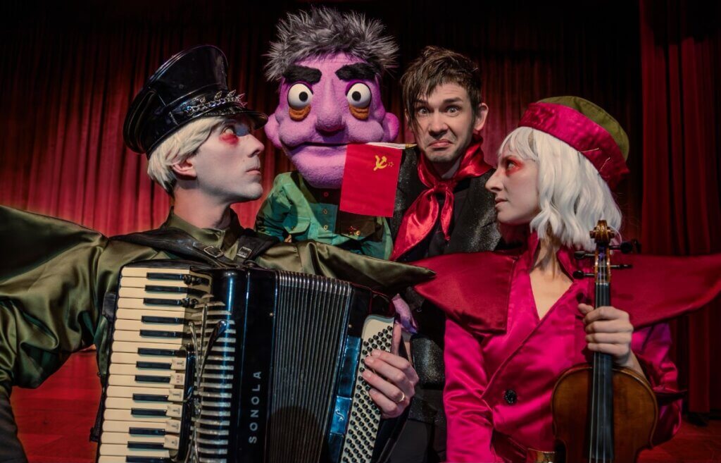 A man in white face make-up with rouge eyeshadows, wearing a Soviet hat, exaggerated green jacket, holding an accordion, stares at an oversized, purple, child-like big-headed puppet Comrade Bucket that embodies a Soviet military person, who is grumpy, tired and strict, symbol and is holding a flag of USSR in their mouth. A woman matches has white make-up with rouge eyeshadow and wears a shade of red jacket and is holding a violin. Next to Comrade Bucket is Timur, who is wearing a black shirt with a red young pioneer tie around his neck. Timur looks scared and confused.