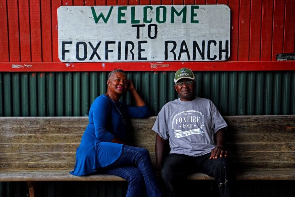 A dark-skinned woman and man, husband and wife, sit on a rustic church pew in front of a wall painted a vibrant red on the top half, and covered with forest green corrugated tin at the bottom. Above them, a white, hand-painted sign with green and black letters reads, “Welcome to Foxfire Ranch.” Her legs are crossed and she wears a blue shirt and jeans, her elbow rests atop the pew, her hand behind her head. He wears a green baseball cap with Mississippi on it in white lettering and a heather gray t-shirt with the Foxfire Ranch logo.