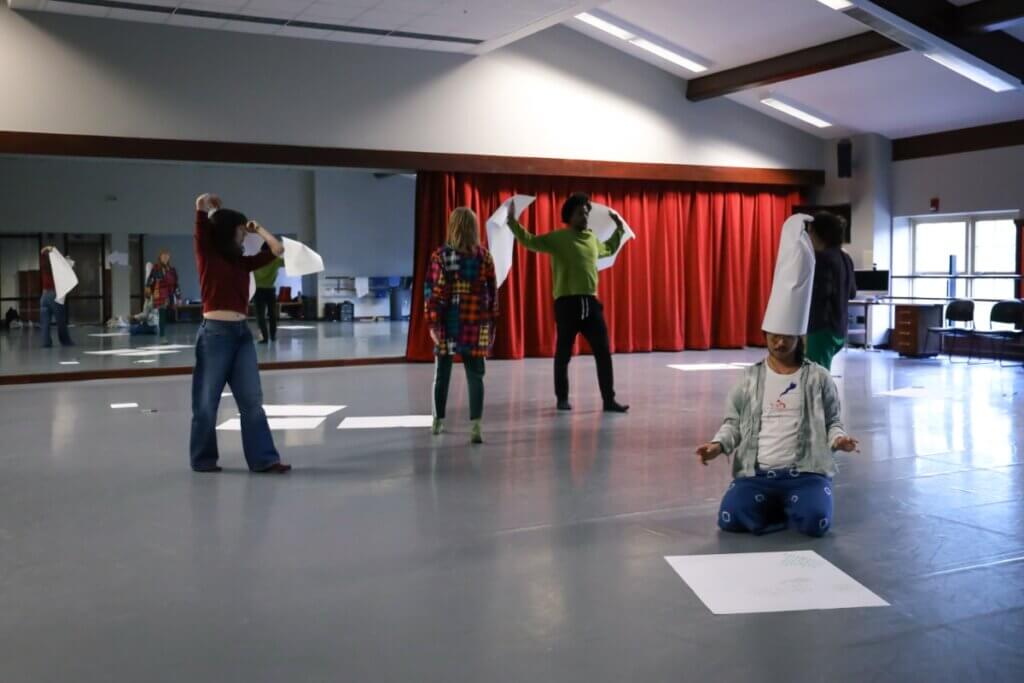 Five dance-makers of various complexions and ethnicities engaged in a collective process. They wear loose clothes and socks. The dance studio has one mirrored wall, half-covered by a red curtain. Huge pieces of paper are scattered on the floor. Two artists are mid-stride. Another stands with two pages aloft like wings. Another has flipped a page overhead like a cape. In the foreground, the fifth artist kneels in front of a page marked with writing in multiple colors and orientations – eyes downcast, hands poised as though about to conduct an orchestra – wearing a piece of paper like a hat.