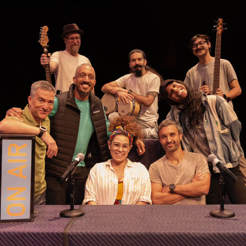 A group of people of various ethnicities and genders pose behind a low, wide table. On the left side of the image, a sign that reads "ON AIR" is placed vertically, and one of the men in the group is resting his arm on it. A woman and another man are sitting behind two microphones in the center of the image. Behind them are two other company members smiling with heads tilted, and behind them are three men holding, left to right, a guitar, a small set of drums, and another guitar.