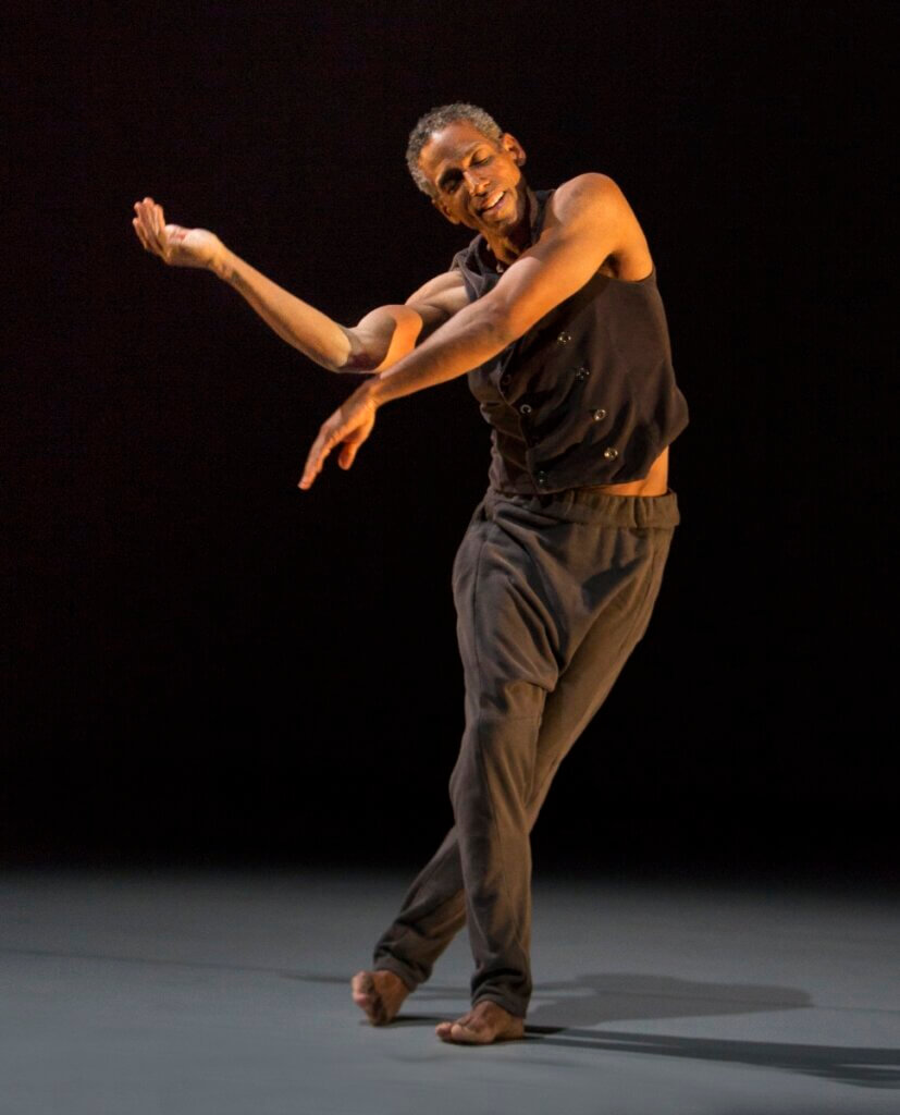 An African American man with salt and pepper hair in his early 60s appears in a standing arc to the right as his bare arms extend rightward in a gesture of reaching and release. He is wearing a black, sleeveless vest and soft thick black pants, and his feet are bare. As he leans, his back foot rolls onto its edge, creating a sense of soft counterbalance. His clean-shaven face expresses a quiet joyfulness, as if recalling a cherished memory.