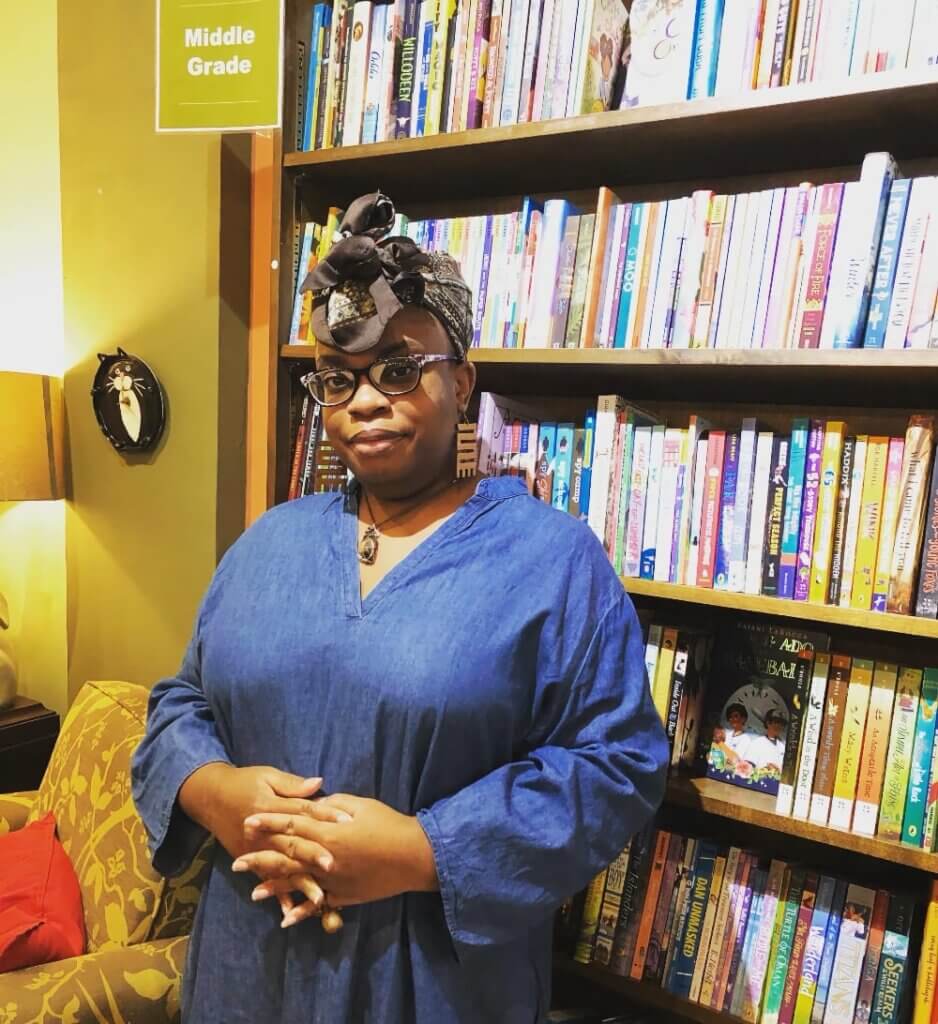 An image of a medium-brown-skinned Black woman standing by a bookshelf smiles at the camera. She is wearing black-framed eyeglasses, a long denim shirt, large brown spiral earrings, a necklace with a crystal, and a brown-and-white patterned head wrap.