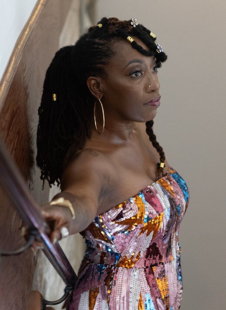 This is an image of a medium-dark-skinned Black woman standing in a stairwell with her back to the wall, looking away from the viewer with her arm extended on the railing. Her loc'd hair is in a ponytail. She is wearing a strapless, multi-colored sequin jumpsuit.