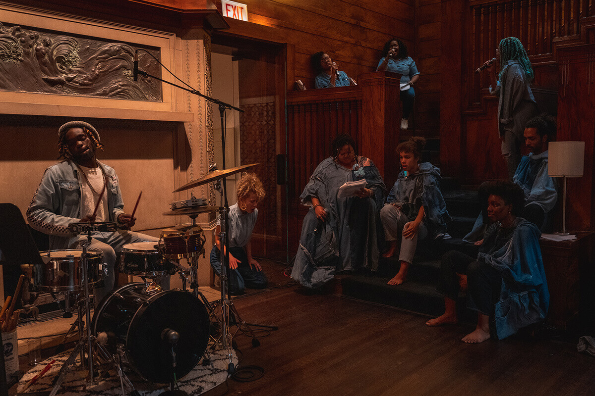A large group of Black folks of all shades and genders—most of them femme—wears denim. They are in a honey oak colored room, taken up largely by a staircase, where some of them sit, some stand and sing. One of them—in the foreground, behind a drum kit—closes his eyes and looks over his right shoulder as he plays drums.