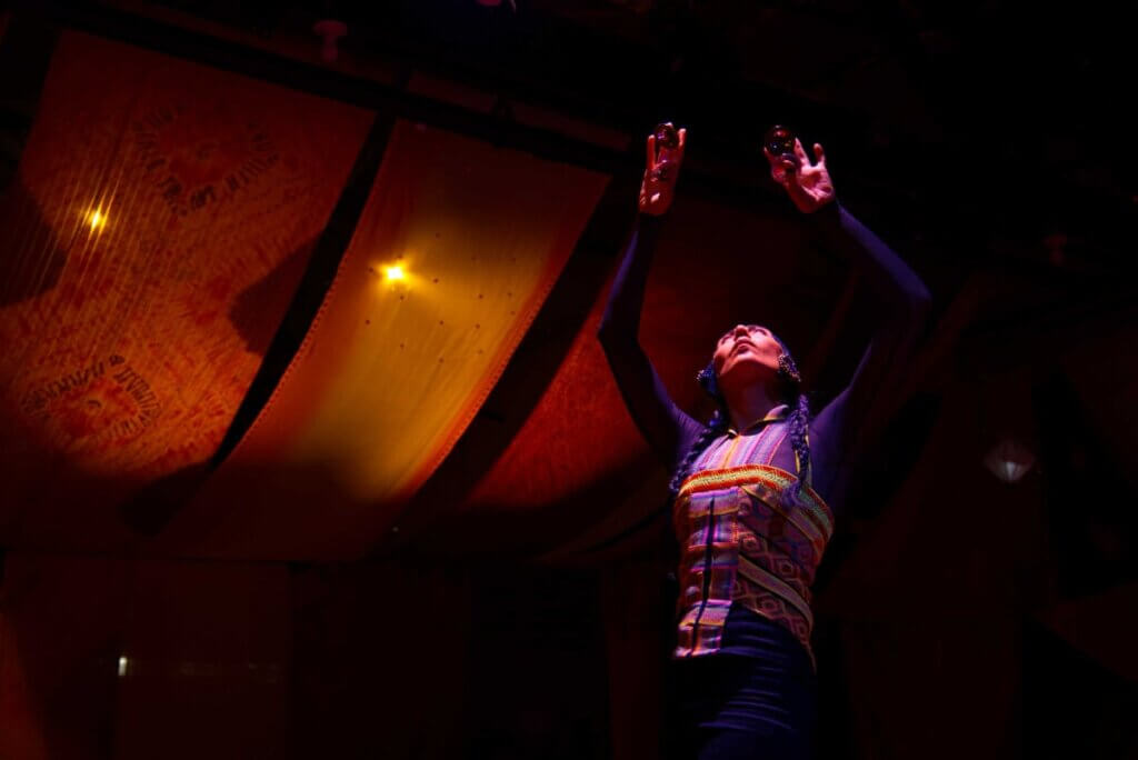 A dancer standing and reaching arms upwards in colorful top with colorful fabric in the back hanging from the ceiling.