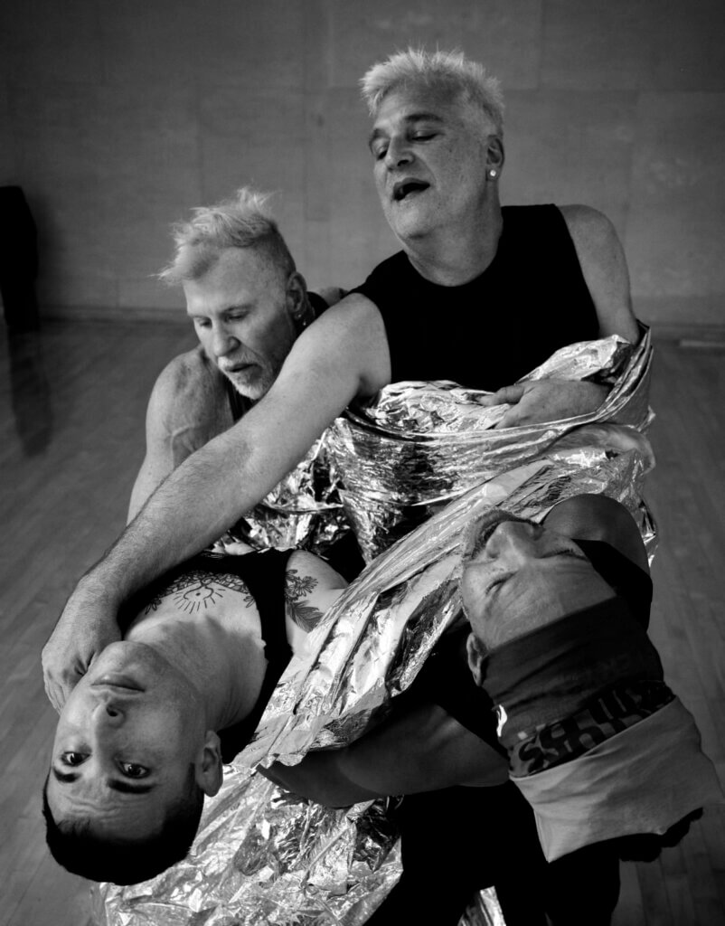 Four dancers in dramatic counter-balance, connected by reflective fabric: Pioneer Winter, a 36-year-old white genderqueer with chest tattoos, arches back alongside Clarence Brooks, a 64-year-old Black genderqueer with dreadlocks. They are supported by Cuban-American 59-year-old Octavio Campos with silver hair, and 66-year-old white Frank Campisano, also with short silver hair.