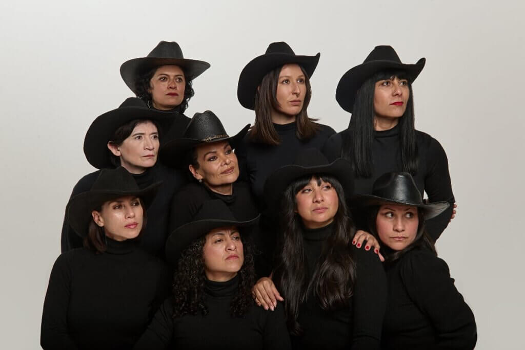 Nine Mexican women dressed in black, wearing black cowboy hats, huddle together and lean on each other, gazing off camera to the right, with serious expressions, and serious attitude.