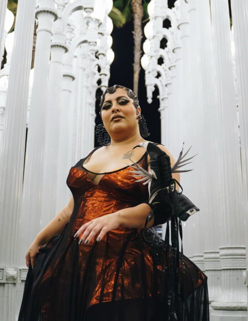 A medium-skinned Latinx performer stands in front of white columns. They wear a dark amber dress, fitted with a corset. On the performer’s left arm, they showcase a bird puppet with silver feathers. Their hair is in an updo with waves, gold and black eye makeup, and large black earrings.