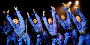 Six Black femmes are positioned in a staggered line. They wear dark blue tracksuits with fringe across the chest and a single shortened pant leg. They all stand with their feet shoulder width apart, hip popped out, and arms stretched above their heads with their torsos leaned away from the popped hip. They stare out in a large stage.