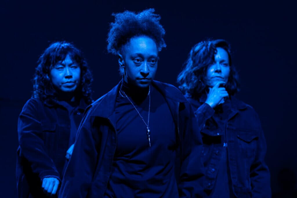 Three femme identified dancers are framed from the waist up in this photo with blue theater light illuminating their skin. They are close together, looking in the distance wearing black jackets, and shirts, exploring the performance of masculine qualities. The dancer to the left is Asian with a wavy dark bob and bangs and a look of defiance. The center dancer is mixed race, black and white, with a high natural poofy bun, a long gold necklace, and a clear look of determination on her face. The dancer on the right is a white woman with dark wavy bob and long bangs swooped to the side. She is holding her hand to her chin, as if she is skeptical of the situation.