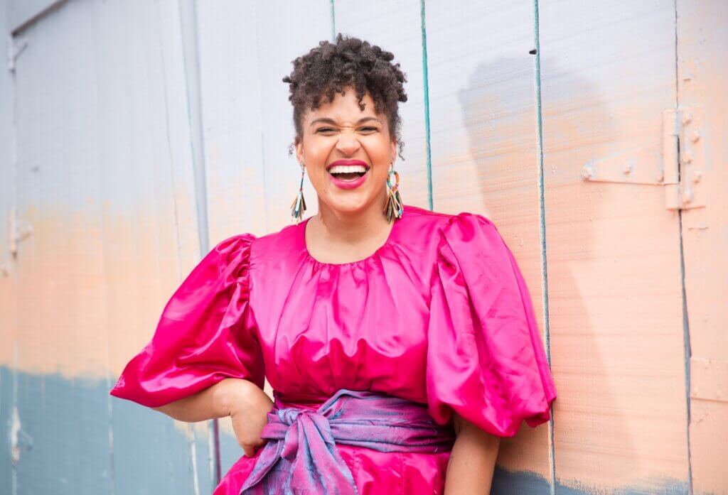 Photo of a lively, smiling Black American woman wearing a voluminous pink dress, curls about her face, and her hand on her hip.