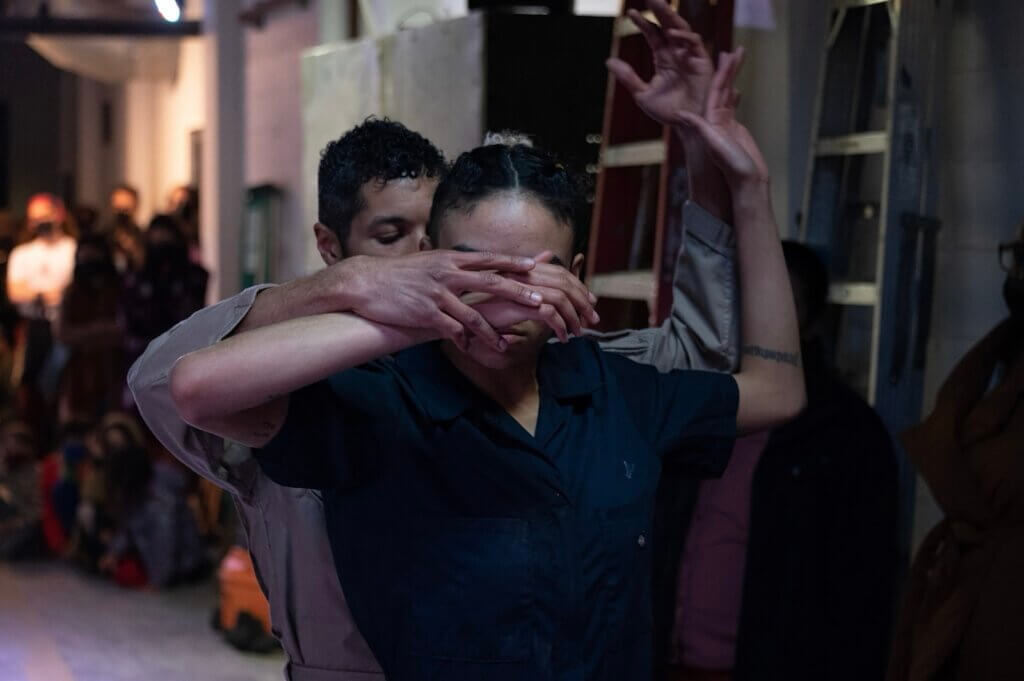 A mid-waist image of two medium-tone brown skinned people, one standing behind the other. The face of the one standing in front is covered by their own right hand and the right hand of the person behind. Both their left hands are extending upwards in unison. They are in a basement hall space. Blurred in the background are audience onlookers gathered against the walls.