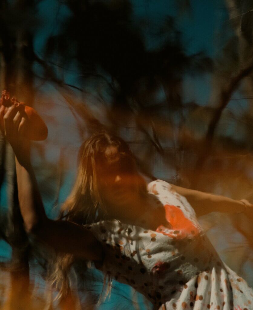 The image portrays a woman standing in a forest, immersed in a dreamlike and distorted atmosphere. She wears a light, polka-dotted dress stained with vivid red marks, and warm orange flares pass across the frame, creating a surreal, almost burning haze. Her posture is slightly tilted, her head lifted as if searching for air or light, giving the moment an emotional tension between fragility and revelation. In her hand, she holds what appears to be her own heart—an intensely red, organic form that contrasts with the soft tones of her dress and skin. This gesture adds a raw symbolic weight, evoking themes of vulnerability, sacrifice, and exposure. Her expression is distant, contemplative, as though she is confronting a truth pulled out from within herself. Behind her, dark silhouettes of trees cut through a deep blue sky, grounding the scene in nature while also amplifying its haunting, otherworldly quality. The entire composition feels suspended between reality and hallucination, as if capturing the precise instant when emotion becomes physical and the inner world breaks through the surface.