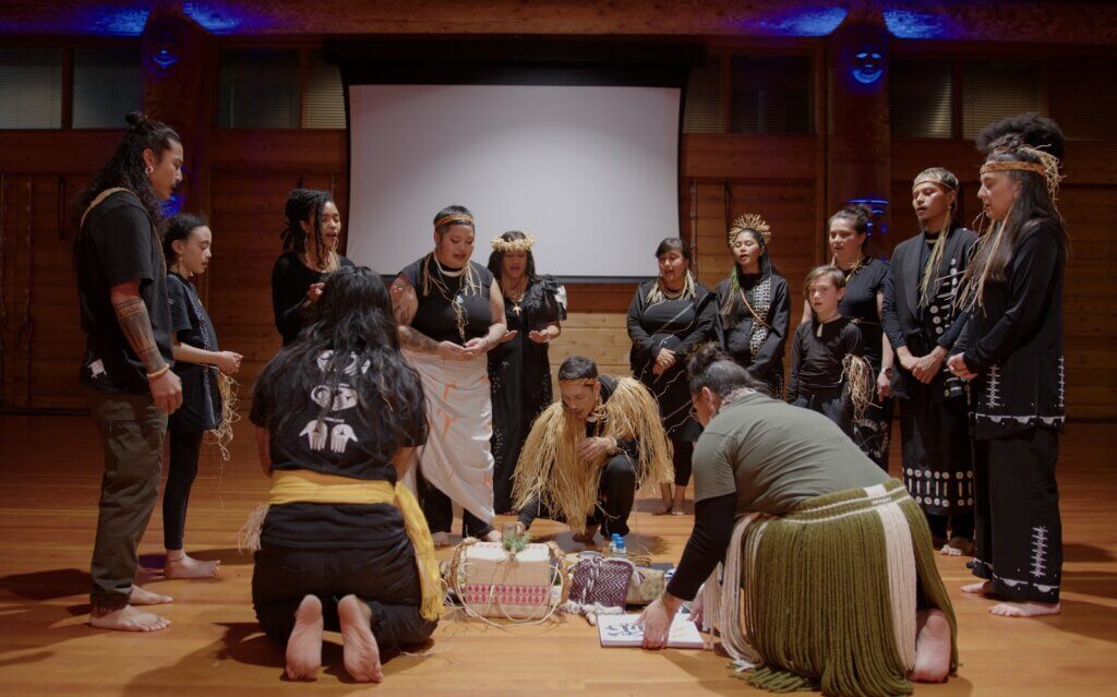 A group of Brown and Black people gather in a half moon around an altar where sacred items are being placed. The people appear to have been gathering in order to create the altar. They are mostly adorned in black clothing with white ancestral designs contrasting their clothes. One of them is wearing all green and a wool skirt. The people are adorned in natural fibers around their heads, ears, shoulders, and necks. Their mouths are open, chanting in unison, some have their hands at their sides, others have their hands raised, and three of them are kneeling down arranging the items in particular order. The group is multi-generational, of different hues, and they stand in a cedar long house with the shadows of statues holding space for them from above.