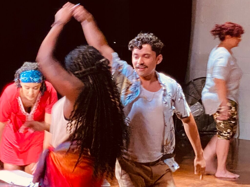 A Latinx man is partner-dancing with a Black woman. We see them from the torso up. He is wearing a floral-printed button-down shirt and a white undershirt, facing us, and she is facing away from us, spinning under his arm. She has long braids and is wearing a dress, with a cream-colored top and a red bottom. A medium-skinned woman in a red dress is in the background.
