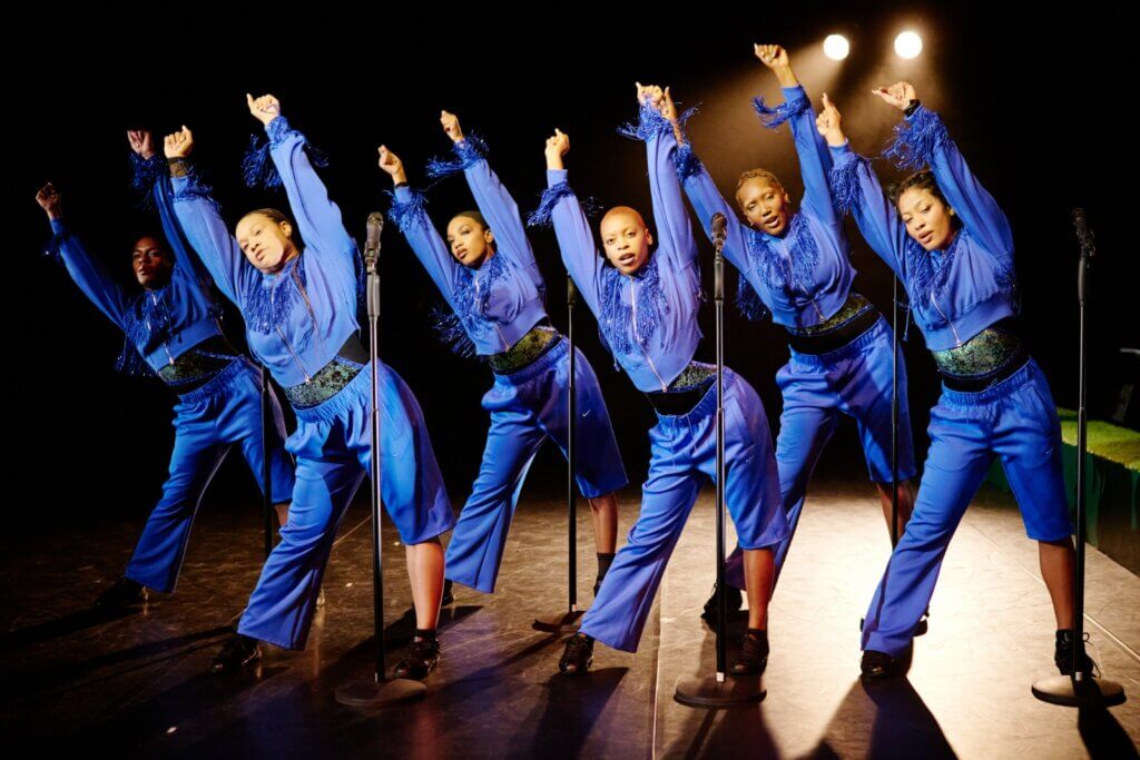 Six Black femmes are positioned in a staggered line. They wear dark blue tracksuits with fringe across the chest and a single shortened pant leg. They all stand with their feet shoulder width apart, hip popped out, and arms stretched above their heads with their torsos leaned away from the popped hip. They stare out in a large stage.