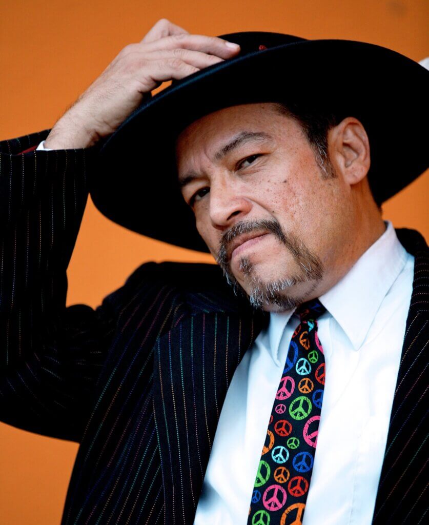 A medium skinned Chicano man dressed in a pin-striped suit, with a white shirt and a multi-colored tie that features round peace symbols. His facial features include a moustache that connects to a goatee. His left hand holds his left lapel, as his right hand reaches for his black, wide-brimmed pork pie Tando (hat), as if about to remove it from his head.