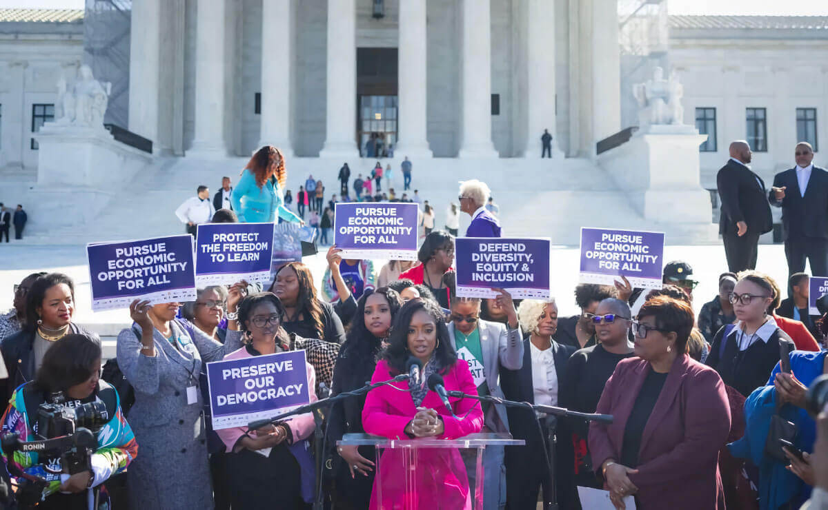 Several dozen women of color wearing business attire stand in front of the United States Supreme Court building. In front of them, a young Black woman wearing a pink coat stands behind a clear speaker's podium and makes an announcement into a pair of microphones. The women behind her are holding up signs with white text on a blue background. The signs read, "Pursue Economic Opportunity for All," "Protect the Freedom to Learn," "Preserve Our Democracy," and "Defend Diversity, Equity & Inclusion." 