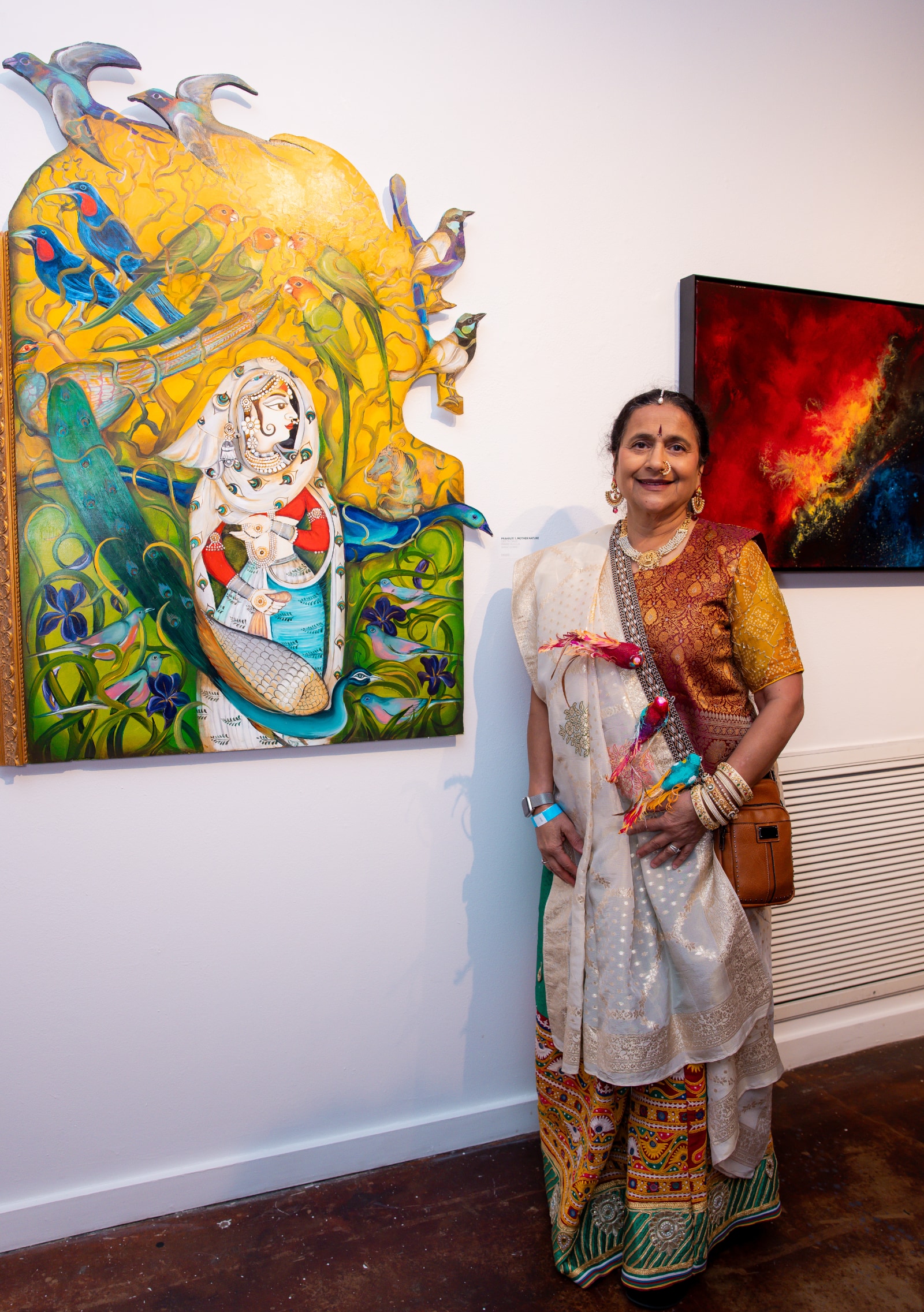An Indian American woman, Geeta Dave, stands in a gallery next to her painting. She is dressed in a traditional Sharara suit in gold and red, with intricate green and yellow patterns on the bottom half of the suit. She has a dupatta of pale peach with gold embroidery draped over her right shoulder. Three realistic looking life size parrot figures, with red, blue, and gold feathers, are fastened to her dupatta. Her painting features a colorful scene of harmony between nature and a woman, with blue flowers, green vines, a distant horse, three peacocks, a bright yellow sky, and various other types of colorful birds in pairs. A segment of decorative gold frame runs along the left side of the painting, while the top and upper right section of the painting has been cut away so that four of the birds extend beyond the yellow sky and appear to be escaping the physical boundary of the work.