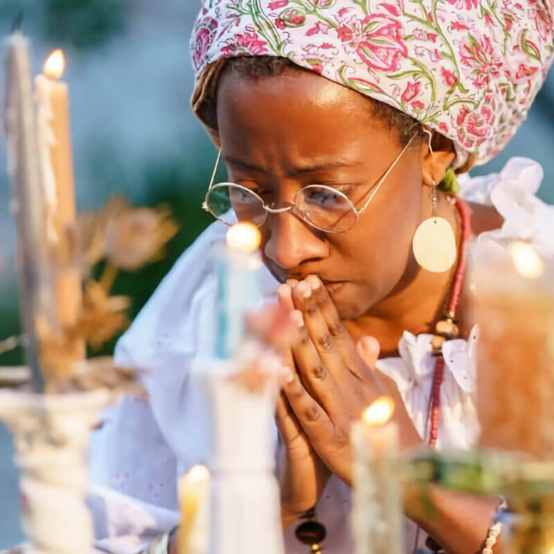 A medium-brown skinned woman in a tignon (head wrap) and wire rimmed glasses, praying at an altar made up of lit candles of different heights. The woman is clasping her hands flat together in prayer, eyes closes, and is wearing a white blouse, red necklace, and shiny round earrings.