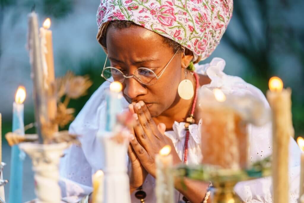 A medium-brown skinned woman in a tignon (head wrap) and wire rimmed glasses, praying at an altar made up of lit candles of different heights. The woman is clasping her hands flat together in prayer, eyes closes, and is wearing a white blouse, red necklace, and shiny round earrings.