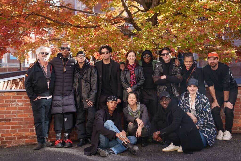 A group photo of 13 individuals from different ethnic backgrounds, dressed in predominantly black outdoor clothing, posed in front of a brick wall and fall foliage.