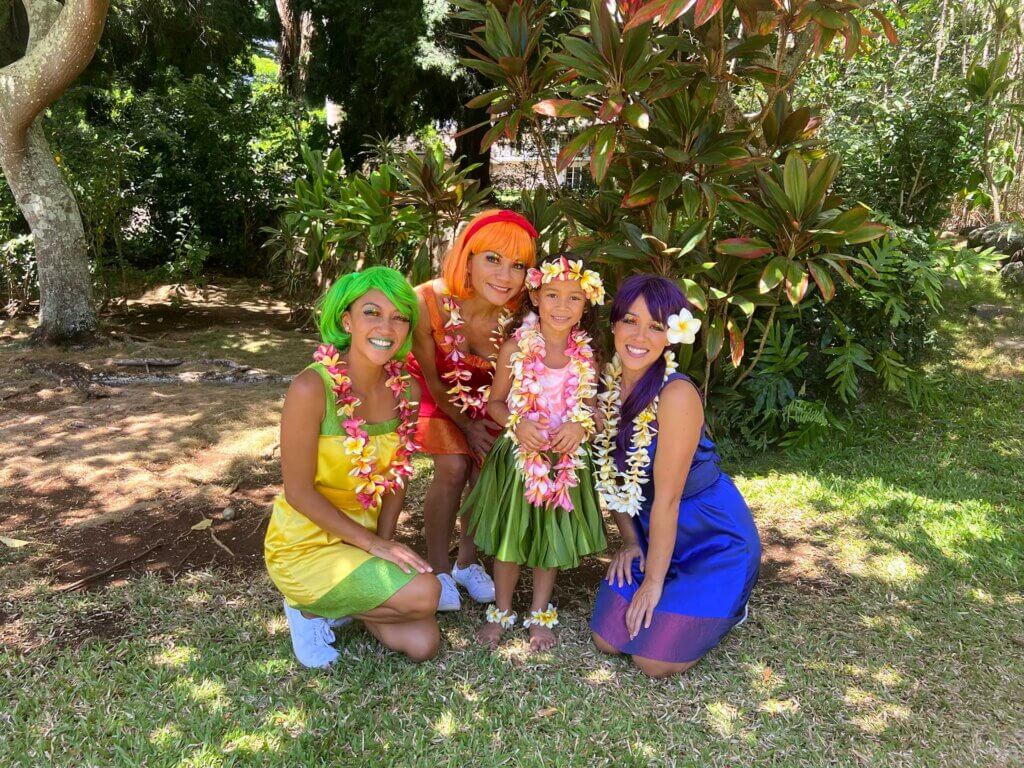 The Peek-a-Bows are three colorful sisters from the rainbow in Hawaiʻi. Lena is dressed in yellow & green with a green wig, ʻAlani is Hawaiian dressed in red & orange with a red wig, & Poni is dressed in purple & blue with a purple wig & flower in her ear. Their young 6-year-old friend wearing a ti-leaf grass skirt. They are all wearing yellow & pink flower leis. They are under the shade of a tree surrounded by green ti leaf plants and lauaʻe bushes. Lena is medium-dark-skinned, ʻAlani is medium-skinned, Poni is light-skinned. Their keiki (child) friend is medium light-skinned.