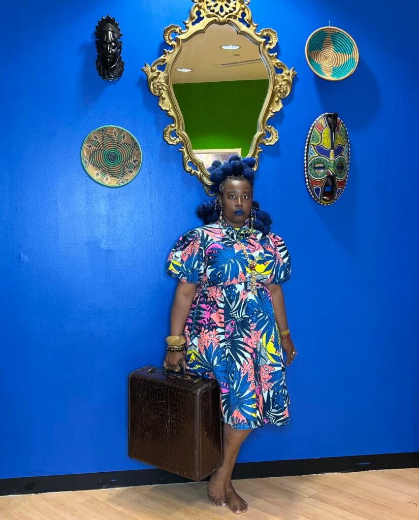 This image shows a dark-skinned, barefoot woman with blue hair styled in tribal braids. She is wearing a blue Ankara dress against an ultramarine blue wall and holding a vintage letter suitcase. African masks, a large mirror, and baskets hang on the wall.