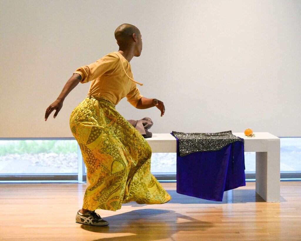 Jasmine Hearn, a person with brown skin and a shaved head, turns their head over their left shoulder looking back towards a white bench. Jasmine is wearing a yellow, orange, and sage green print dress. Draped over the bench is a cobalt blue garment made by Athena Kokoronis of DPA underneath a silver studded black top. An orange citrus fruit is near the edge of the bench. The walls are white, and the floor is wooden.