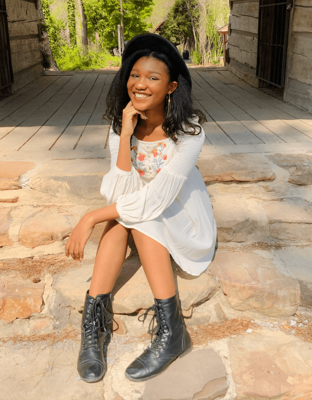 Jazzie Jelks, a young Black woman, sits outside on stone steps at the edge of a covered wooden walkway between two buildings. She is smiling at the camera. She is wearing a white long sleeved dress with embroidered pink and red flowers across the chest, black lace-up boots, and a black hat. She is holding her right hand under her chin while resting the other arm over her knees.