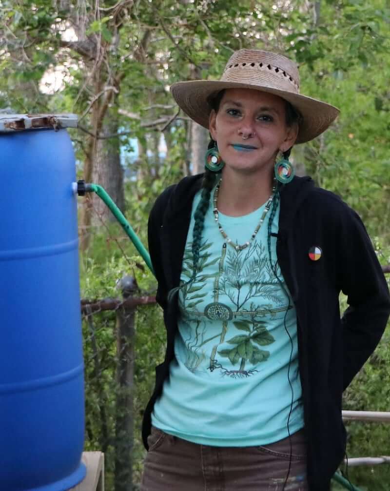 This image is of a light-skinned/tan femme person wearing work pants, a light green shirt, and a straw hat standing outside next to a rain barrel connected to a house's gutter system. They styled their/her dark hair in two long braids, and they are wearing green Indigenous beadwork earrings and bright blue lipstick. Trees are in the background.