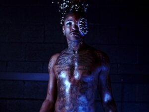 Performer Juicebox P. Burton, visible from the waist up, stands in a dark space, their body illuminated on one side by a cool white light and on the other side by a blue light, and stares intently into the camera. They are unclothed, and tattoos cover their torso, chest, shoulders, and neck. They have blue and silver eye shadow, and the left side of their face has been transformed with prosthetics and makeup into a large, nightmarish circular mouth filled with concentric rows of sharp teeth. Their hair is decorated with what appear to be small silver or white spheres that reflect the light.