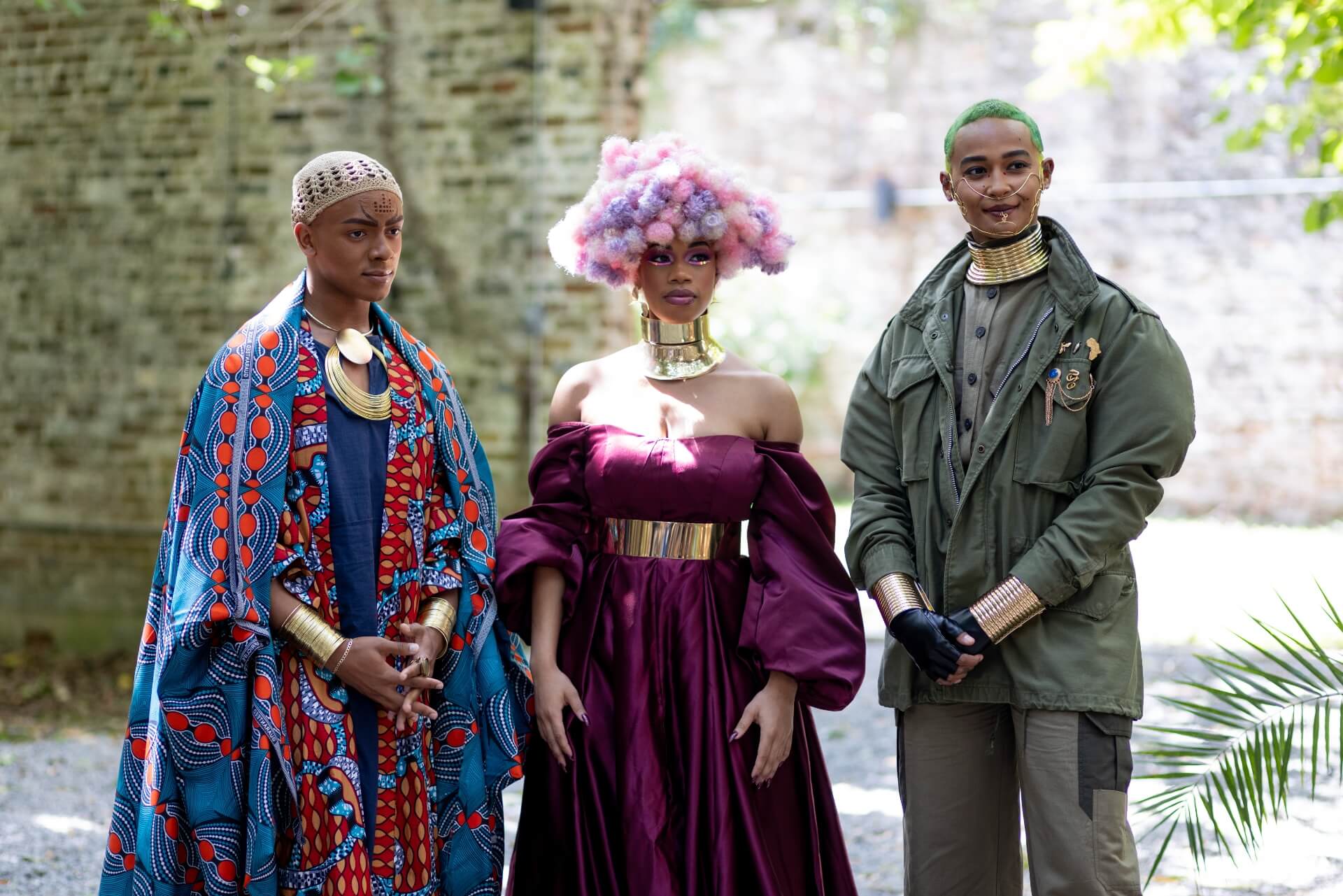 Three actors in the film “Space to Breathe” stand together in a shaded outdoor setting. They are all staring to the right of the camera, as if listening to someone speak. The actor on the left is wearing a blue and red gown, gold wrist bands, a large gold necklace that covers most of the chest, and an ivory colored, knitted skull cap. The actor in the middle wears a voluminous royal purple off-the-shoulder gown with puffy sleeves, a wide silver belt, a silver neck collar, and a wig that appears to be made of small white, pink, and purple spheres of hair. The actor on the right wears sage pants with black utility pockets on the side, a sage high-collared shirt with black buttons, an army green coat with gold pins over the left pocket that include a silhouette of Africa, a gold neck collar, and bronze wrist bracelets. The actor’s hair is closely cropped and bright green, and there is a delicate face decoration made from curving gold wire across the lower half of the actor’s face.