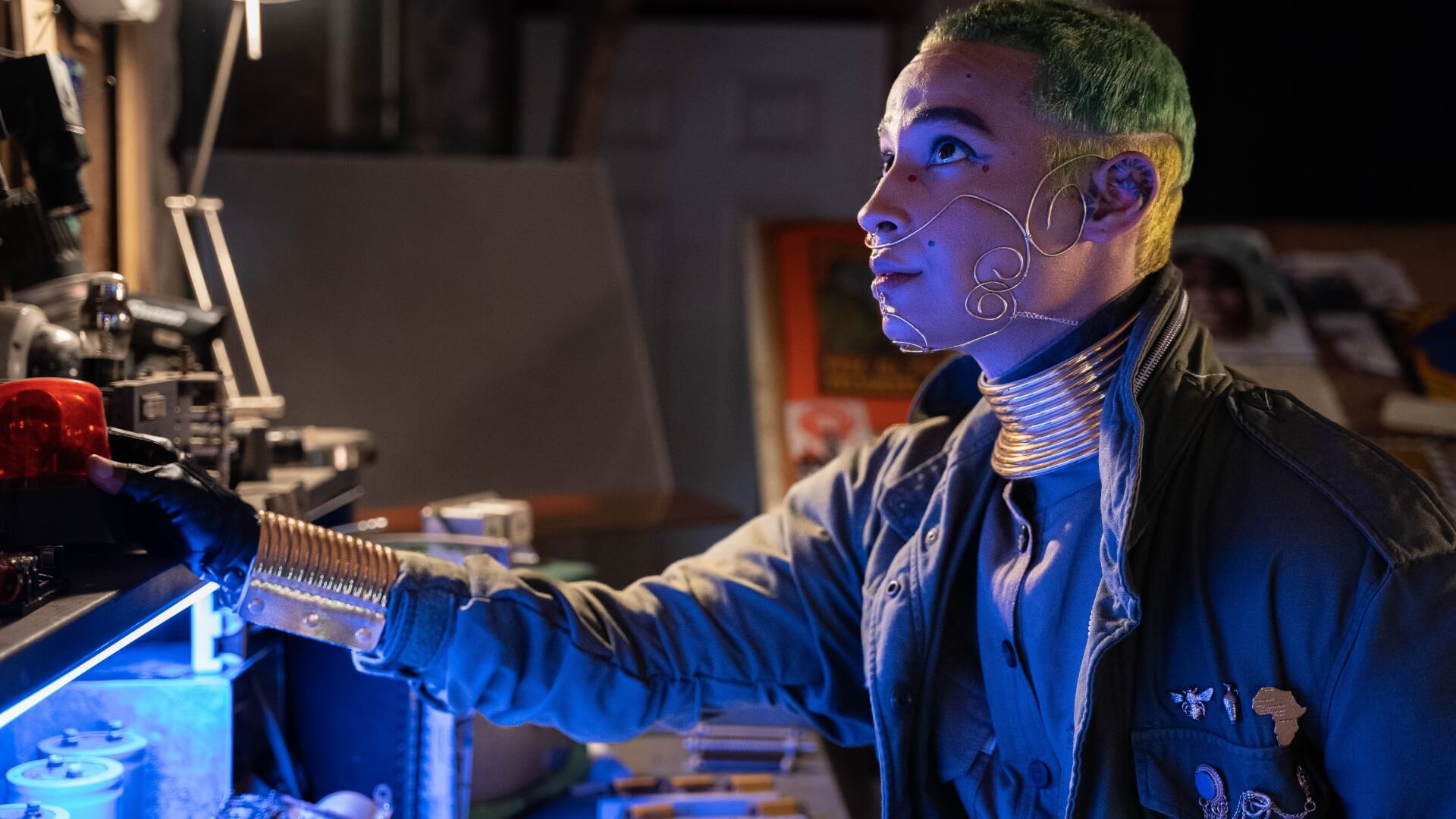 In this still from the film “Space to Breathe,” a character with closely-cropped green hair and a face decoration made from curving gold wire that extends across the lower half of the face, leans forward in a cluttered, darkened workspace and looks up at something out of frame, as blue light from a source below plays across their face. They are wearing a large coat with pins over the left pocket, including a bumble bee and the silhouette of Africa.