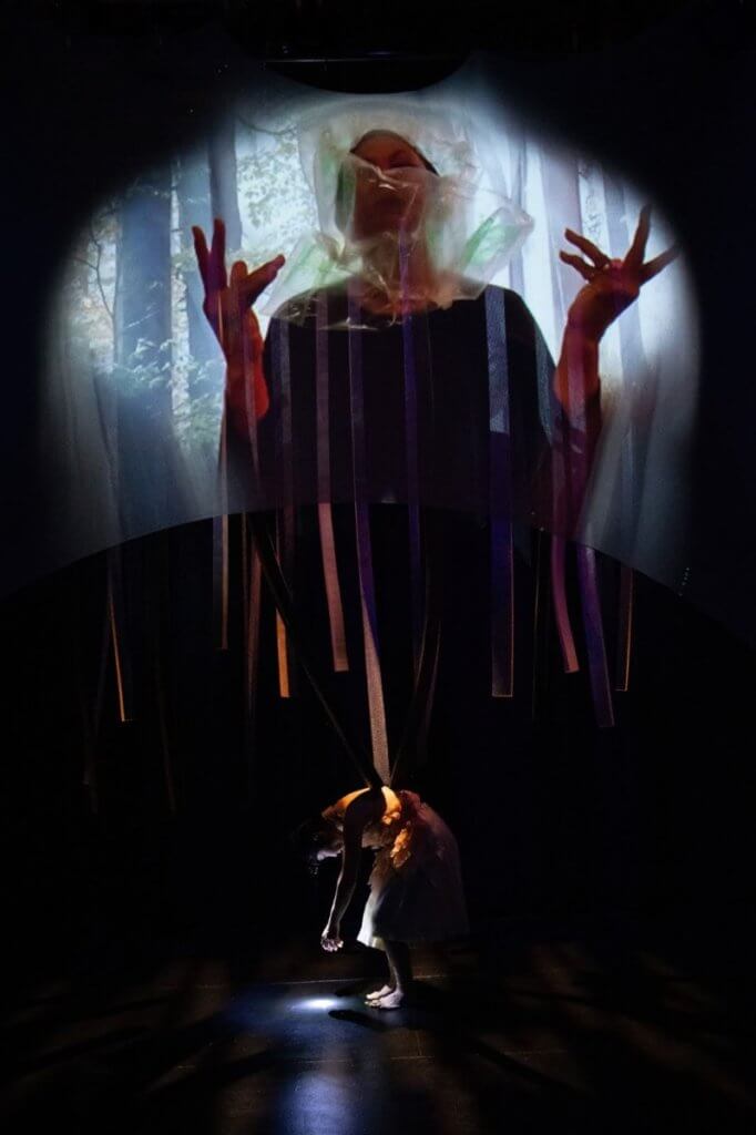A figure projected on a screen above a stage. They are standing in a forest and raise their arms gesturing, while their face is obscured by clear plastic. A figure onstage is bent over, as if in mid-movement as they rise to stand.