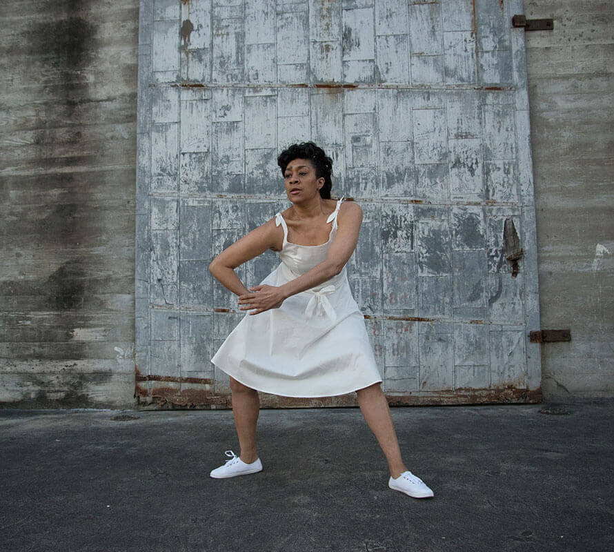 A medium light-skinned African American woman mid-movement dancing in front of a large rustic gray metal panel attached to a concrete wall. Her knees bent in a slight squatting position with arms in a stirring motion crossing in front of the torso at the hands. She is wearing a white knee-length dress that has bows tied at the tops of the shoulders, a white waist belt tied in a bow and white flat string-up sneakers.