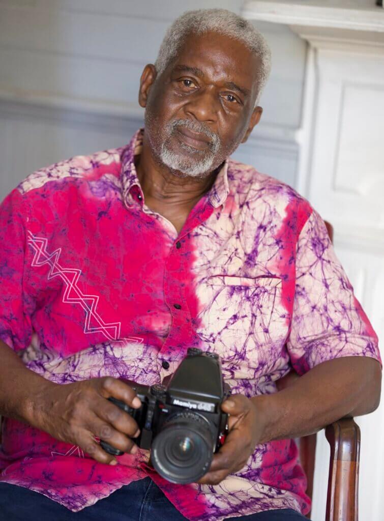 This image shows a dark-skinned African American man with gray hair and a goatee. He is sitting in a wooden chair, holding his camera while looking at the viewer. He is wearing a tie-dyed fuchsia, purple and white button-down shirt and dark blue jeans. The background is a gray wall and a white-painted fireplace mantle.