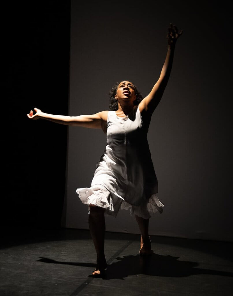 The image of a light-skinned African American woman wearing a loose white knee-length tank dress in mid-movement while dancing under a white spotlight on a black box stage. She is in a lunge position, with her arms up and side. Her gaze is upward, and her hair is in relaxed curls.