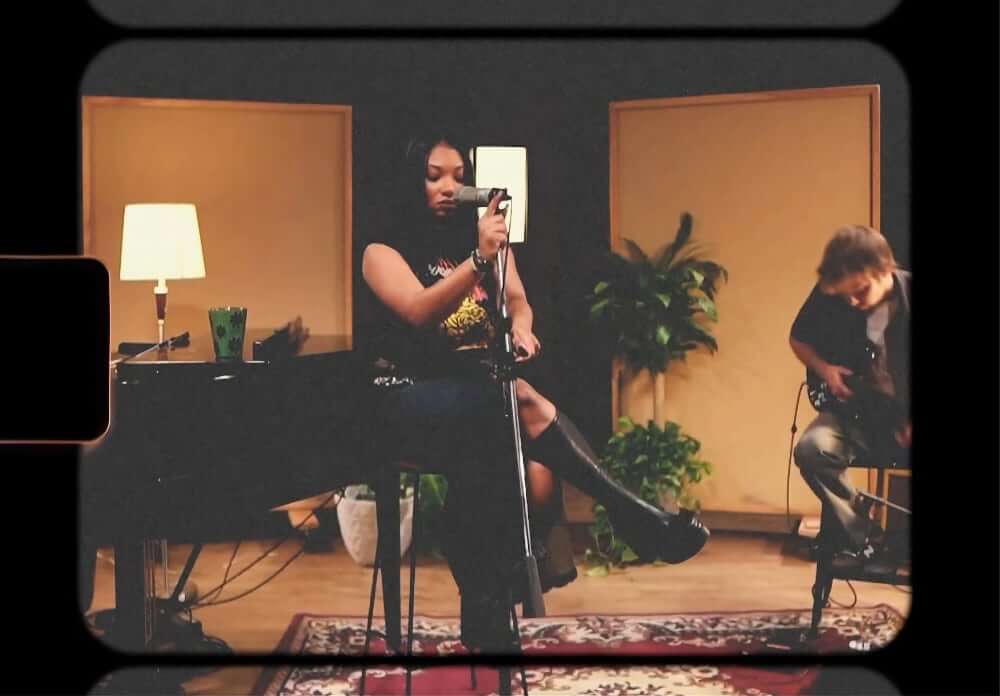 The image of a medium-skinned American American musician sitting cross-legged on a stool while holding a microphone in a dimly lit recording studio. To her left sits a pianist, and to her right is a guitarist. Lamps and plants are in the background, and a cup of tea is atop a black baby grand piano.