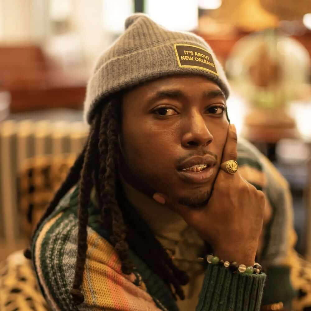 An image of a medium-dark-skinned African American male with a goatee and long locs. He wears a grey knit cap and a multi-colored striped sweater. He looks into the camera, his chin resting in his right hand.