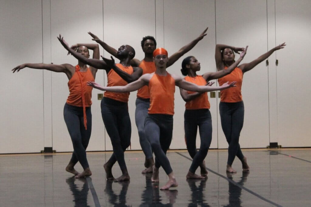 Six Black and Brown performers in orange tops and gray tights pose in a symmetrical vignette against a wall of white doors. Their arms frame each other's faces with an air of balletic formality.