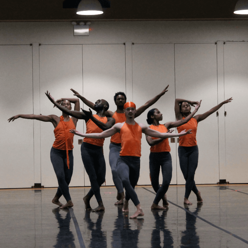 Six Black and Brown performers in orange tops and gray tights pose in a symmetrical vignette against a wall of white doors. Their arms frame each other's faces with an air of balletic formality.