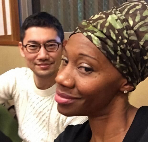 A close up selfie shows a woman wearing a green patterned headwrap and small stud earrings smiling softly toward the camera. Behind her, a man in glasses and a light sweater looks on with a relaxed expression. The warm indoor lighting and tight framing create an intimate, friendly moment between the two.